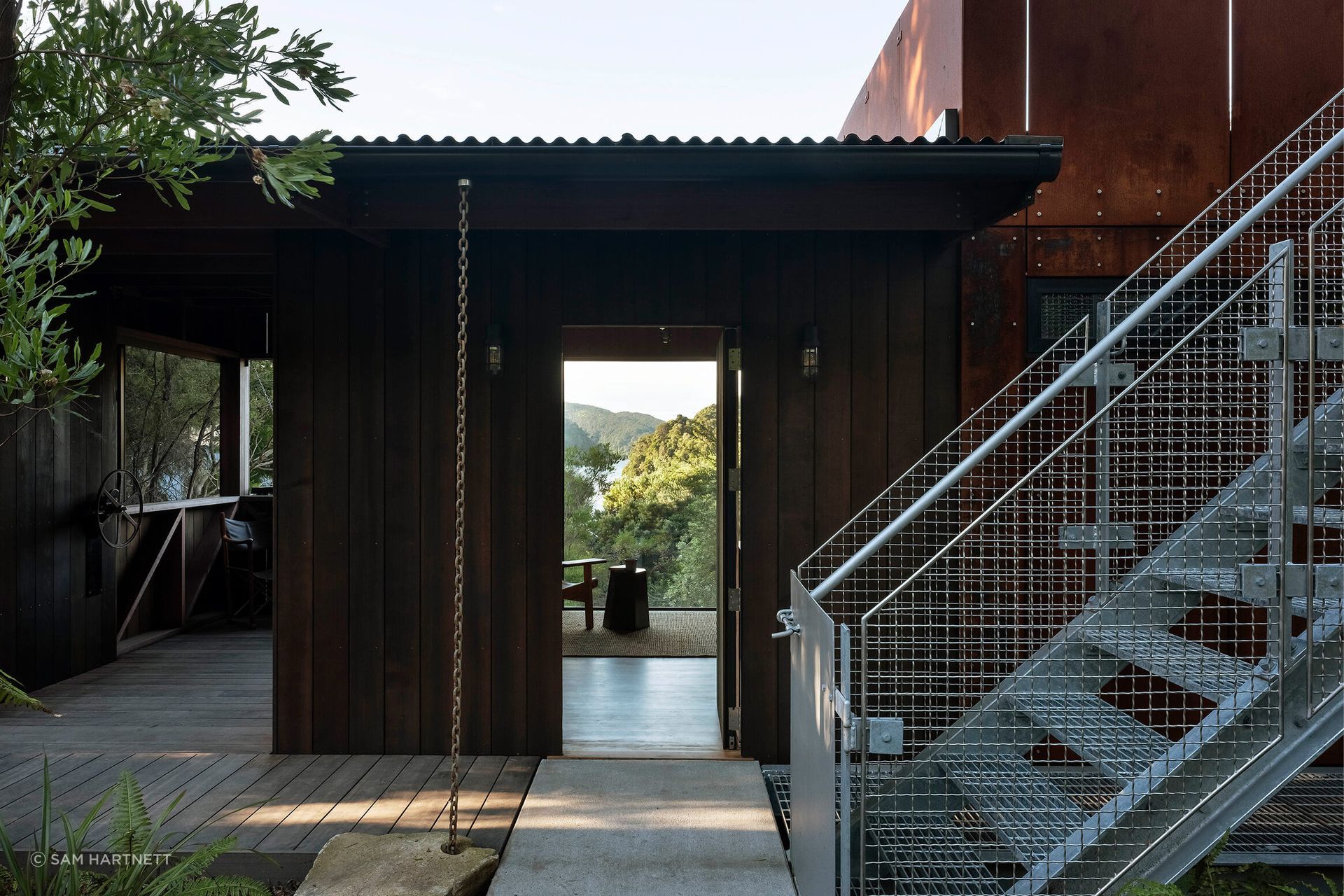A metal staircase climbs to a treetop roof deck where the Cor-Ten cladding extends into a cantilevered balustrade.