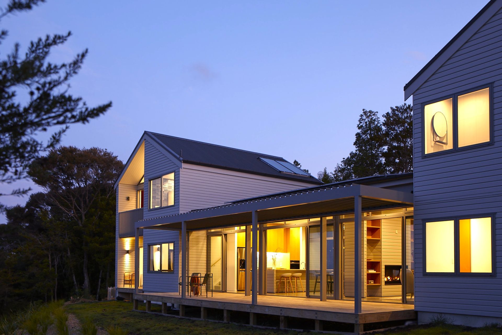 As evening falls, the home glows within its natural setting, revealing the barbell plan that separates the private bedroom wings from the central living spaces. The restrained palette of horizontal weatherboards and corrugated roofing complements the surrounding native landscape.