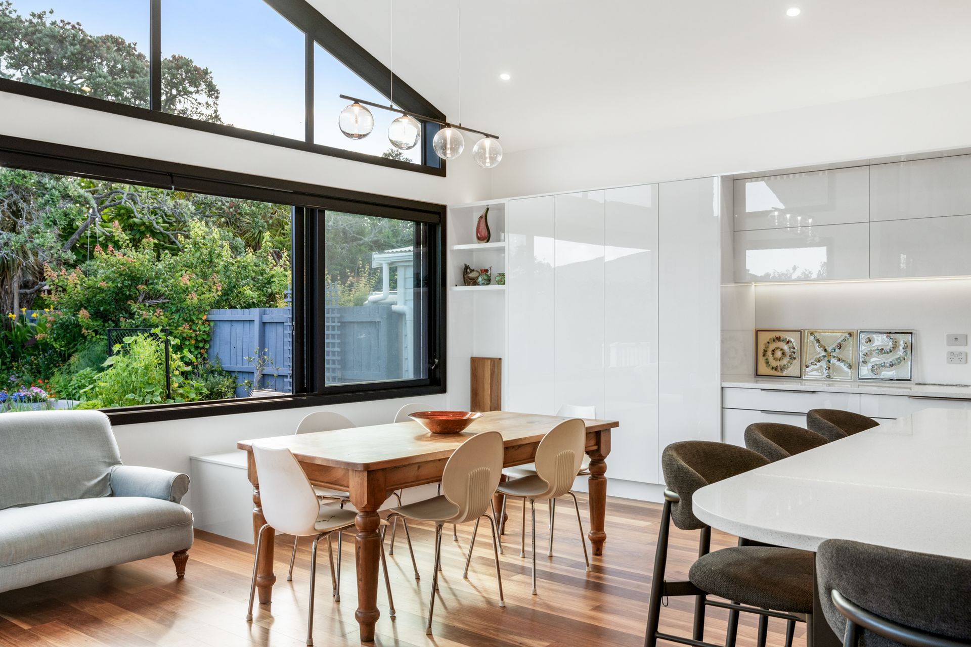 Clerestory glazing framing the garden, globe pendants overhead and a timber dining table at the heart of it — the dining room at Devonport Bungalow. Macfie Architecture.