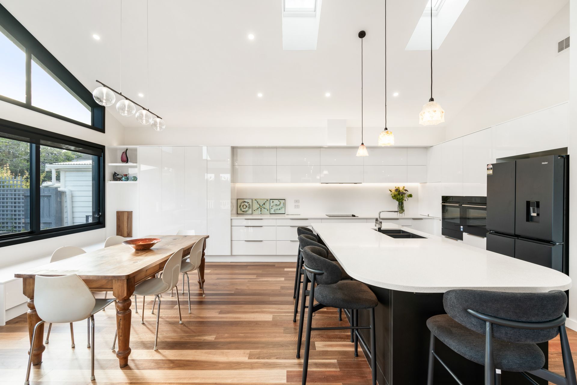 The full sweep of the open-plan kitchen and dining at Devonport Bungalow — gloss white cabinetry, dark island bench and timber floors running through to clerestory glazing and garden beyond. Macfie Architecture.