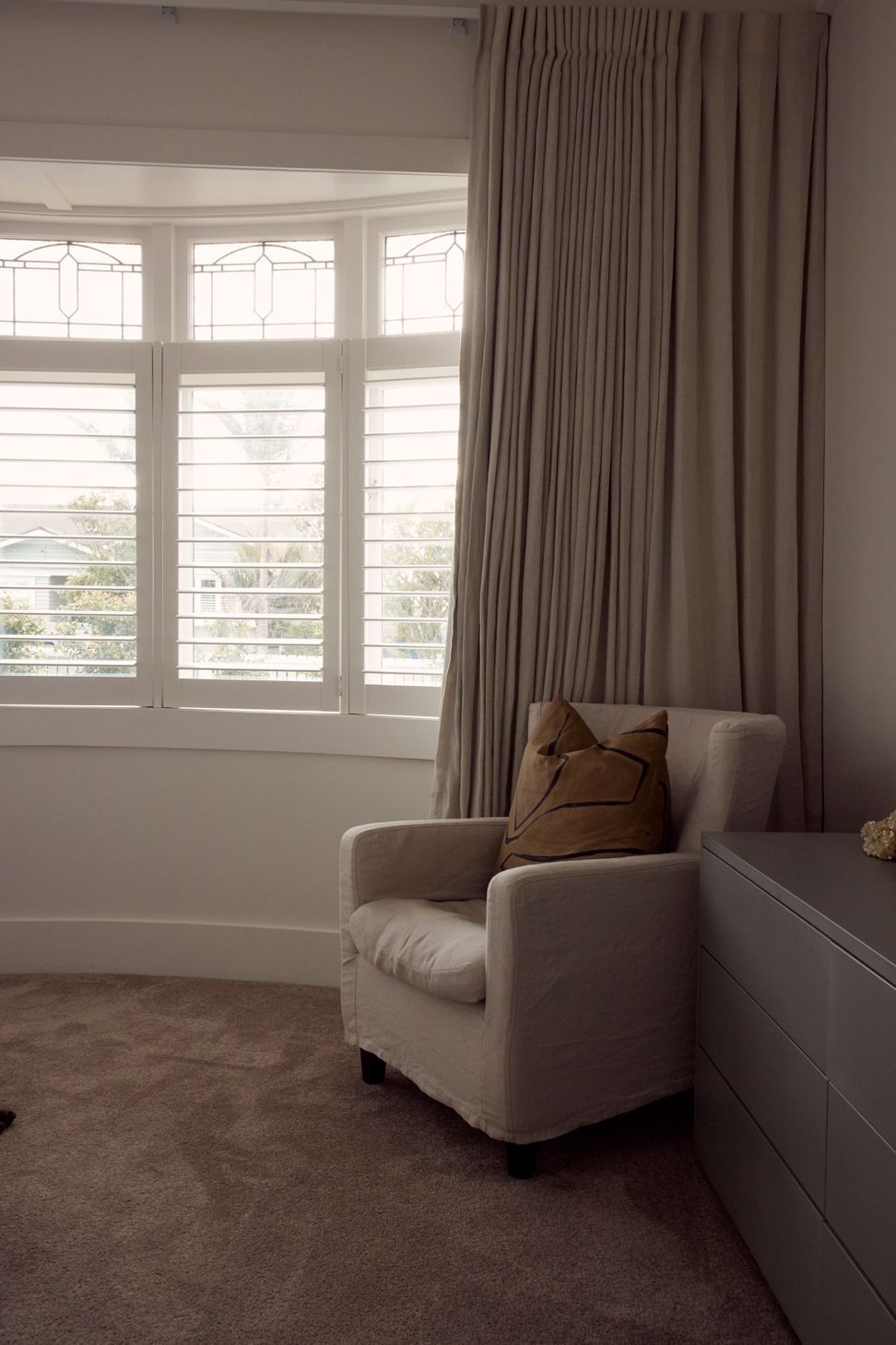The original bay window and leadlight detail retained in the bedroom at Point Chevalier Bungalow — plantation shutters, linen curtains and a considered armchair in the corner. In collaboration with Hannah Warren Interiors.
