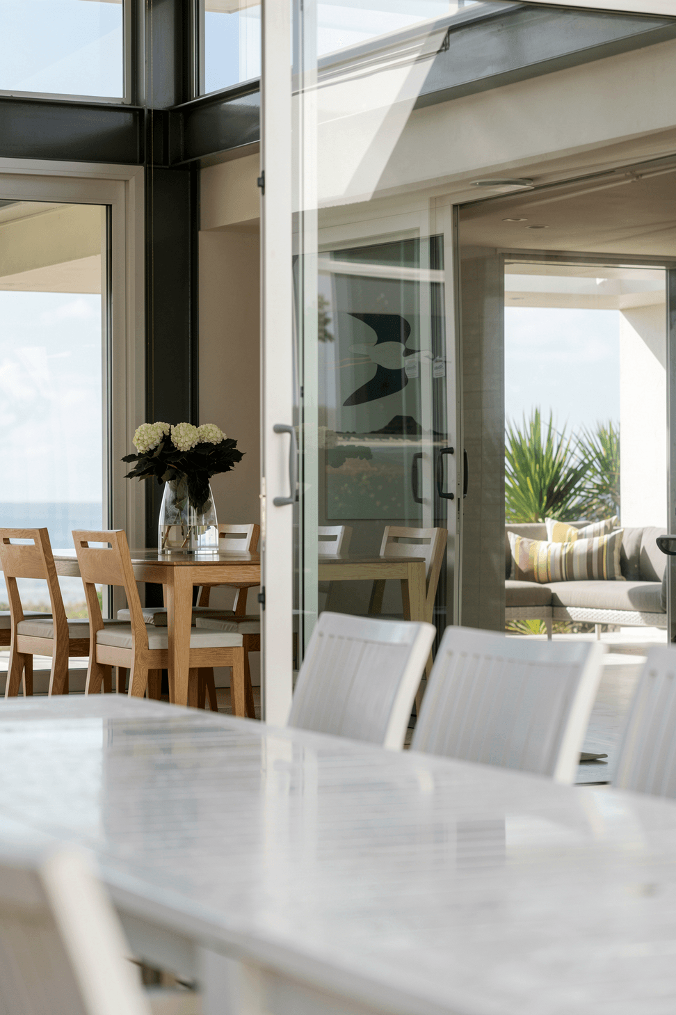 The sheltered internal courtyard provides a calm outdoor dining space protected from the coastal environment. With the large sliding doors open, views extend through the dining room towards the ocean, creating a seamless connection across the home.